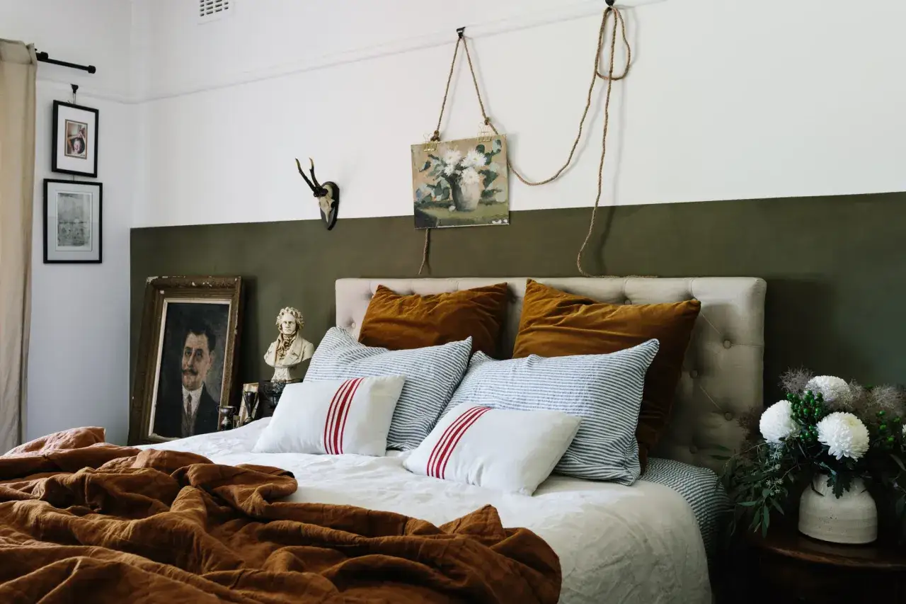 Un letto accogliente con cuscini color terra e a righe, un quadro appeso con corda e un busto. Idee su cosa mettere sopra la testata del letto per un tocco artistico.