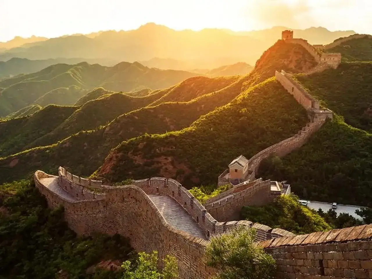 Die Chinesische Mauer schl&auml;ngelt sich durch gr&uuml;ne H&uuml;gel im Sonnenuntergang. Man fragt sich, wie lang ist die Chinesische Mauer wirklich?