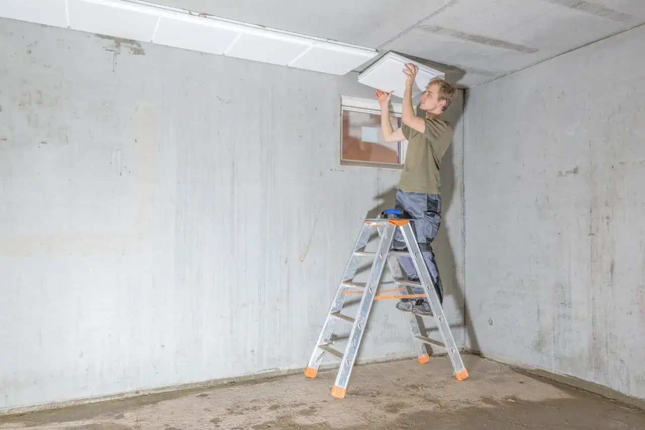 Mann auf Leiter montiert wei&szlig;e Platten an Beton-Kellerdecke zur Isolierung.