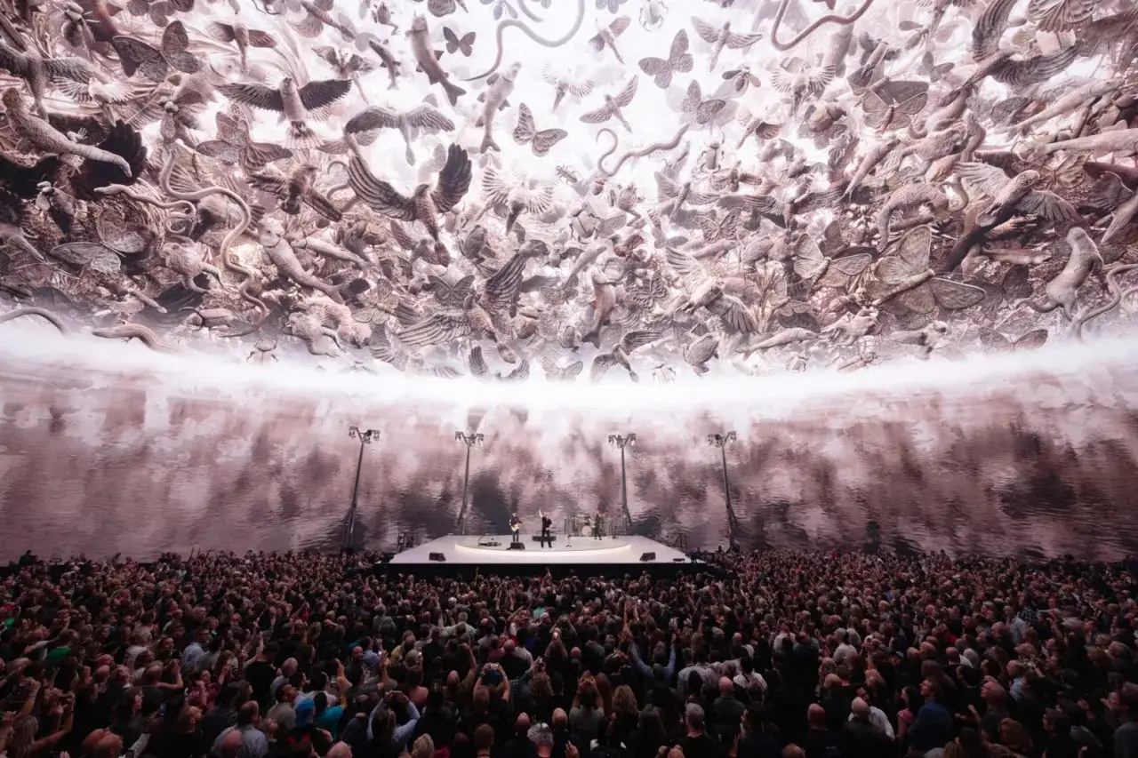 Koncert U2 w Atomic City. Tłum fanów podziwia scenę z zespołem, a nad nimi rozpościera się surrealistyczny sufit z motylami i ptakami.