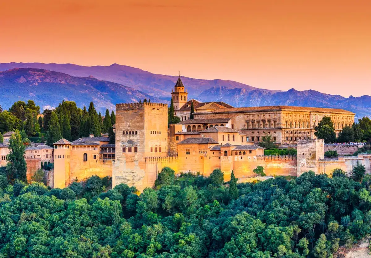 La Alhambra de Granada, un lugar imprescindible que ver en Granada en 3 d&iacute;as, se alza majestuosa entre &aacute;rboles verdes y monta&ntilde;as al atardecer.