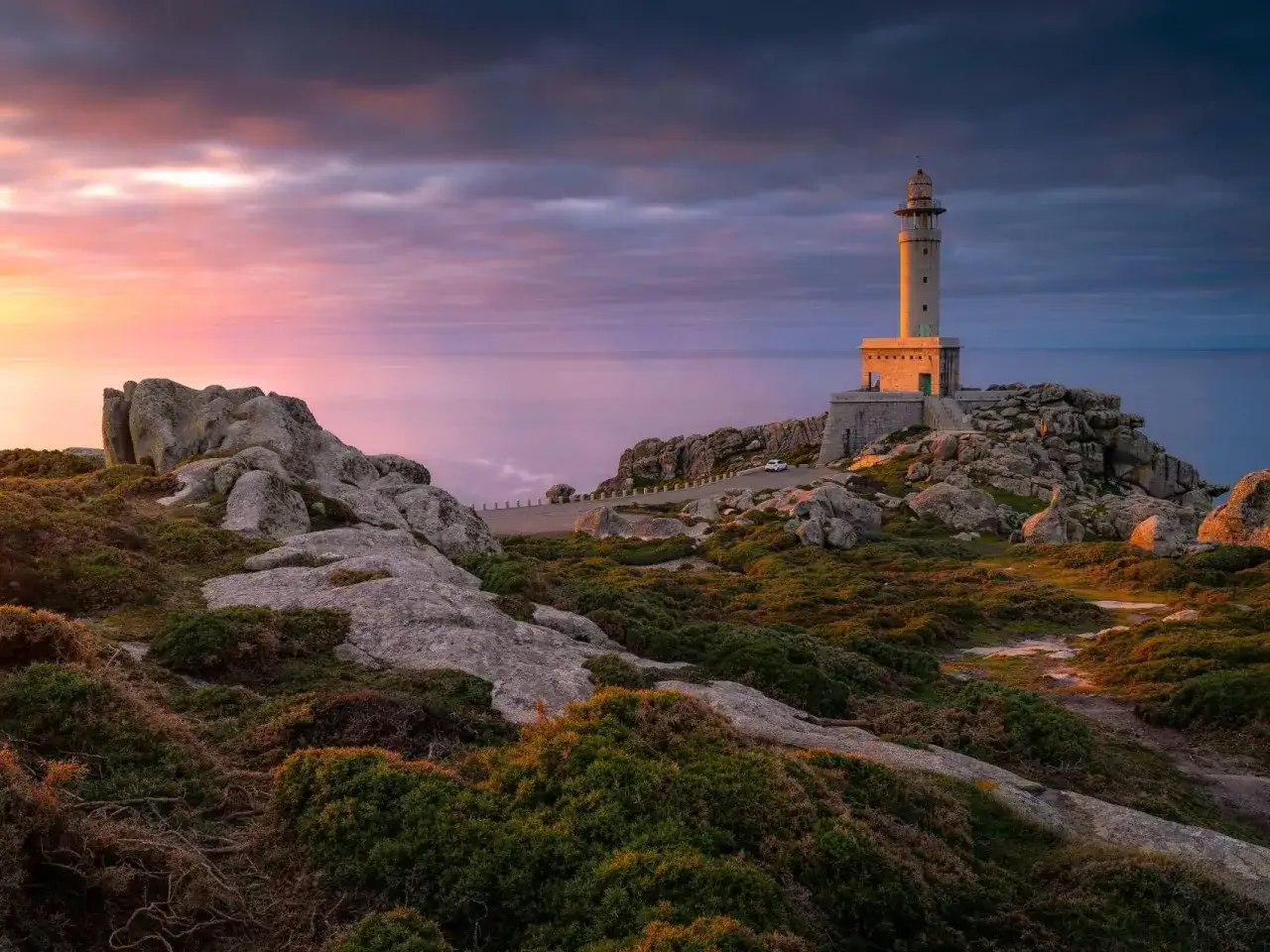 Faro en el cabo Finisterre al atardecer, con el mar en calma y un cielo te&ntilde;ido de rosa y p&uacute;rpura.
