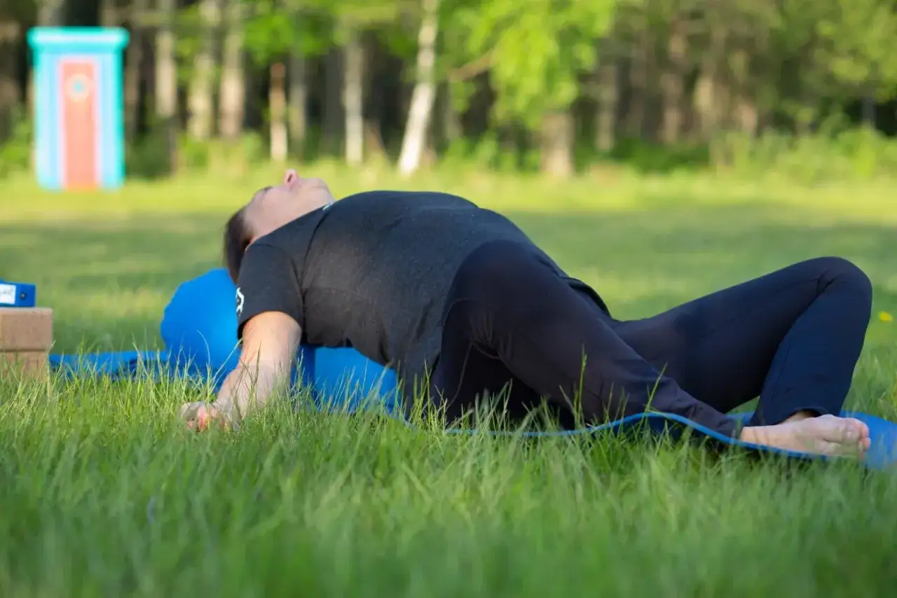 Person utför yin yoga på en blå matta i gräset. En rolig, färgglad bod i bakgrunden. Yin yoga viktminskning kan vara en del av en hälsosam livsstil.