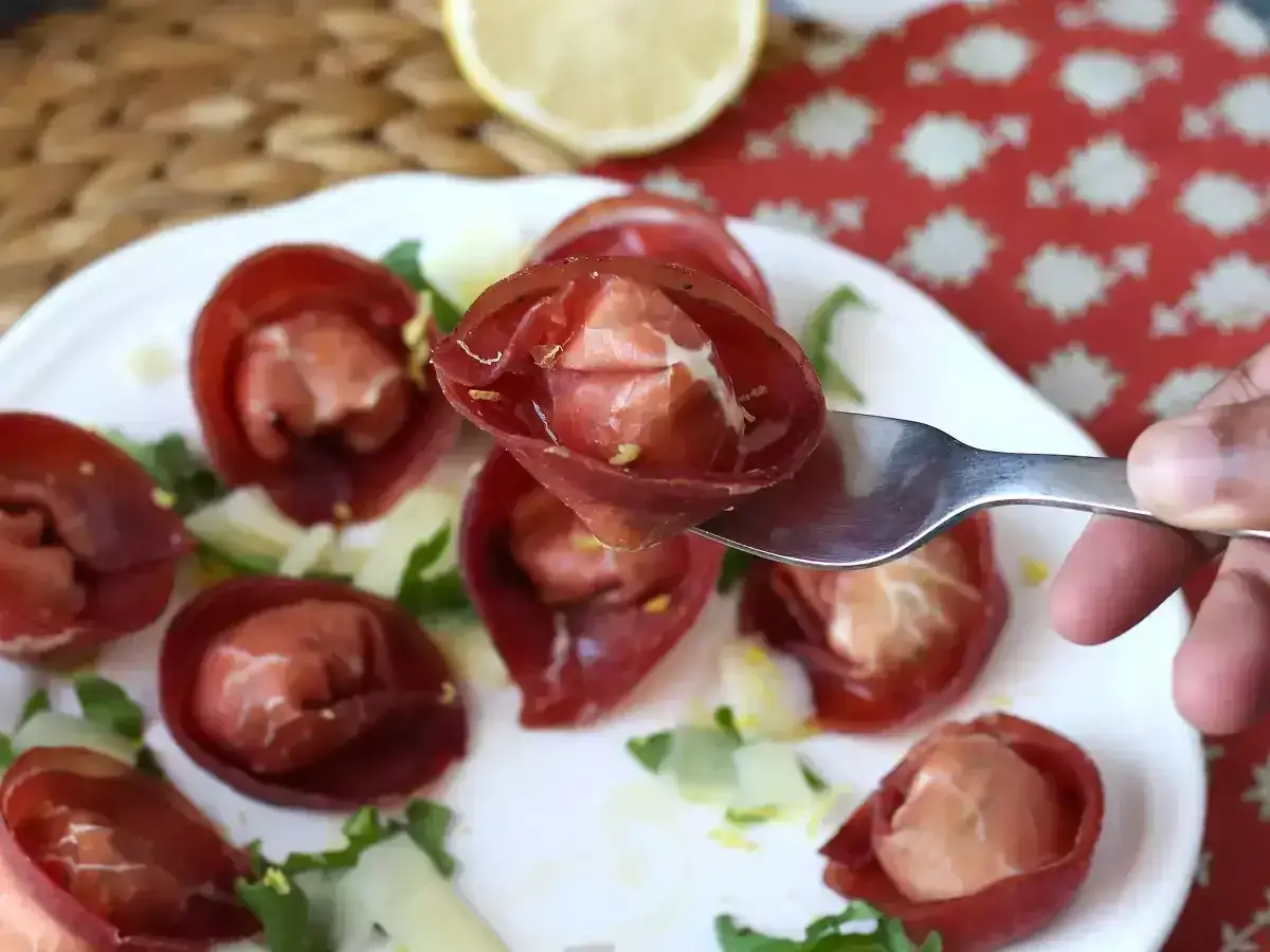 Bresaola co to? Delikatne, czerwone plasterki bresaoli zwinięte w kształt łódeczek, wypełnione serem i rukolą, podane na talerzu z cytryną w tle.