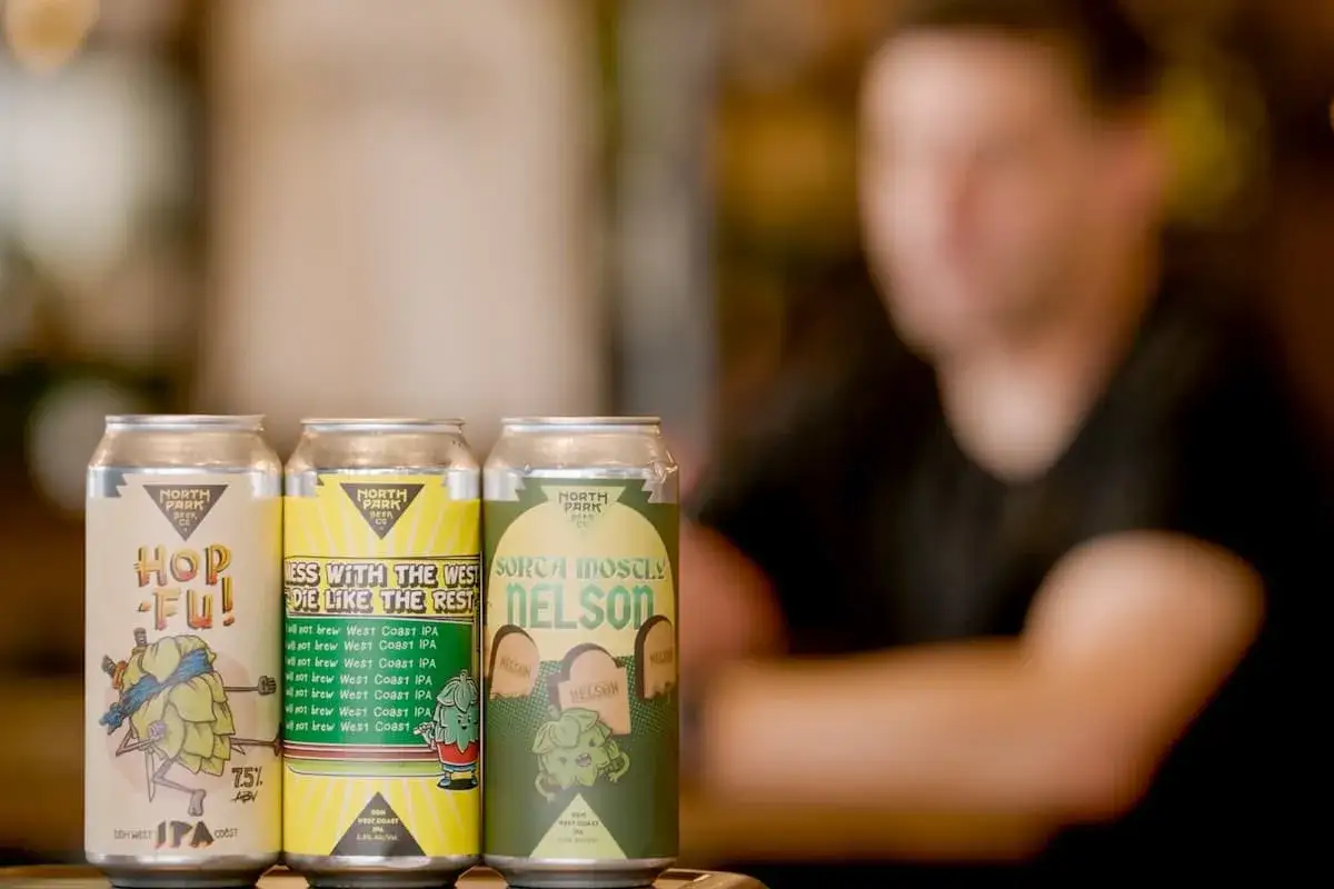 Tre lattine di session IPA della North Park Beer Co. con grafiche vivaci. Una persona sfocata sullo sfondo.