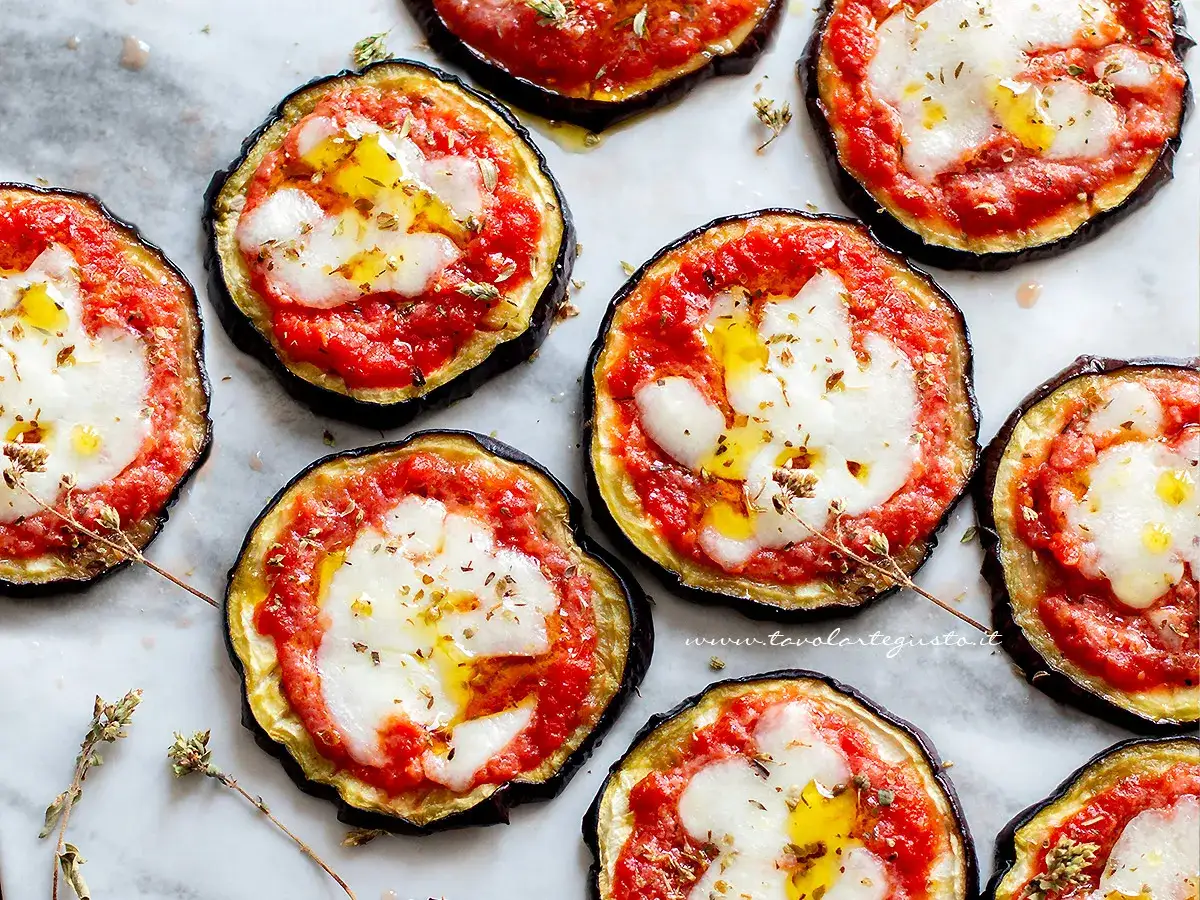 Deliziose pizzette di melanzane con pomodoro e mozzarella filante, condite con olio e erbe aromatiche.