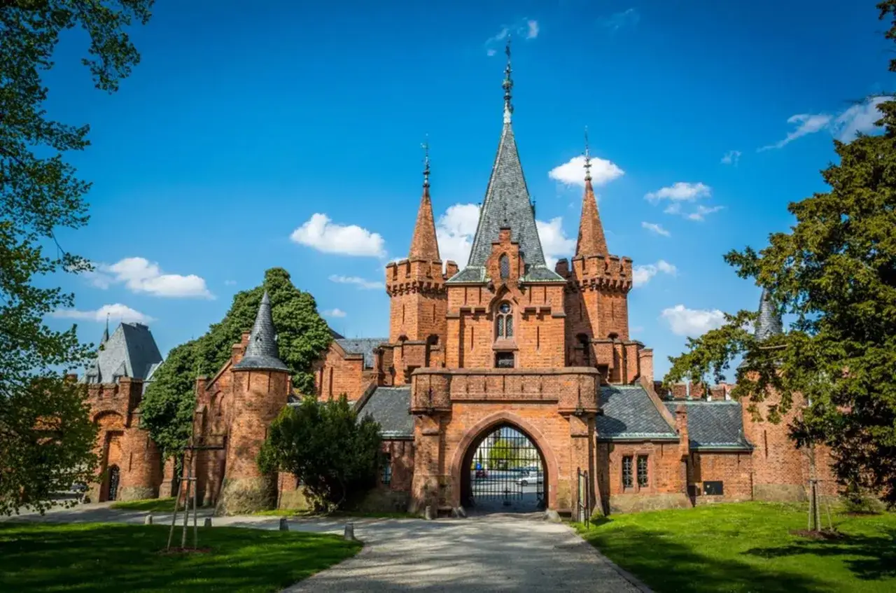 Imponujący czerwony zamek z wieżami i blankami, otoczony zielenią, pod błękitnym niebem z chmurami.