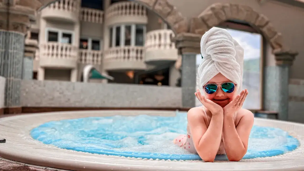 Mała dziewczynka w okularach przeciwsłonecznych i ręczniku na głowie relaksuje się w jacuzzi. Idealne miejsce, gdzie na weekend z niemowlakiem.