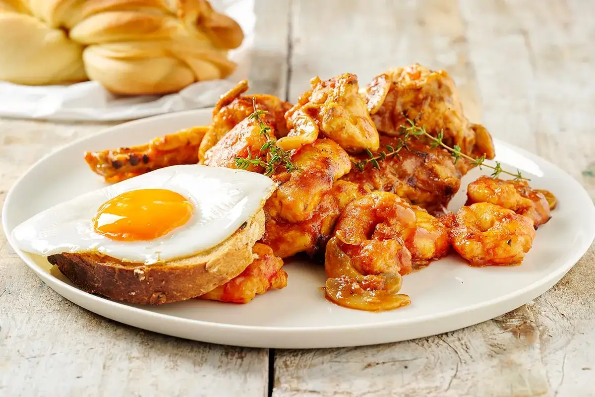 Piatto di pollo alla marengo con gamberi, uovo fritto su pane tostato e un pane intrecciato sullo sfondo.