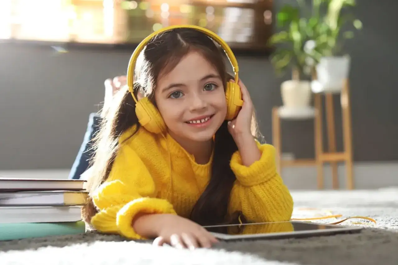 Ein lächelndes Mädchen mit gelben Kopfhörern hört aufmerksam zu. Neben ihr liegen Bücher und ein Tablet – perfekt für **podcast englisch lernen kinder**.