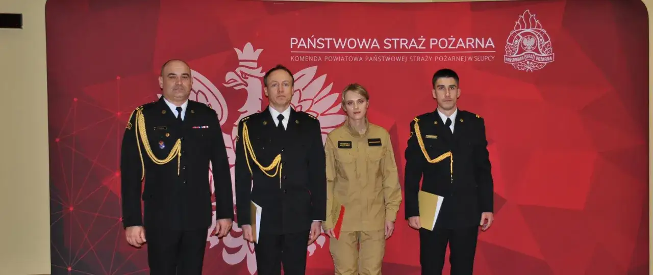 Czterech funkcjonariuszy Państwowej Straży Pożarnej w galowych mundurach i kombinezonie, z odznaczeniami i dokumentami, stoi przed czerwonym tłem z logo.