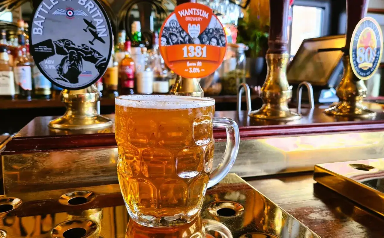 Boccale di birra dorata, con sullo sfondo i spillatori, uno dei quali con l'etichetta 