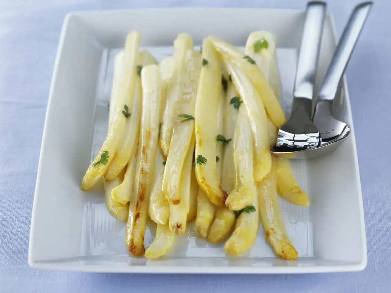 Zarter Spargel in der Pfanne, goldbraun gebraten und mit Petersilie garniert, liegt auf einem wei&szlig;en Teller. Daneben liegen Besteckteile.