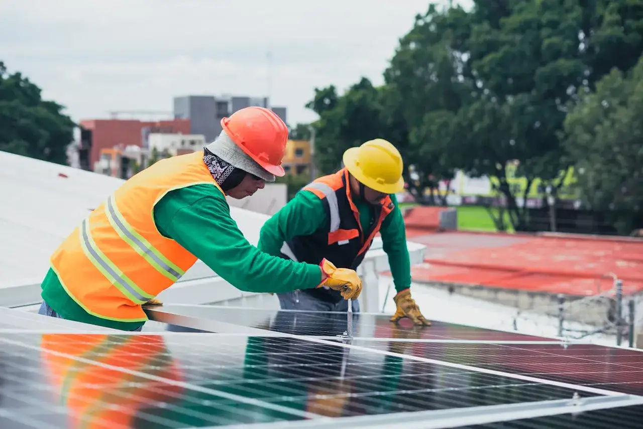 Dwóch elektryków w kaskach i kamizelkach montuje panele słoneczne na dachu.