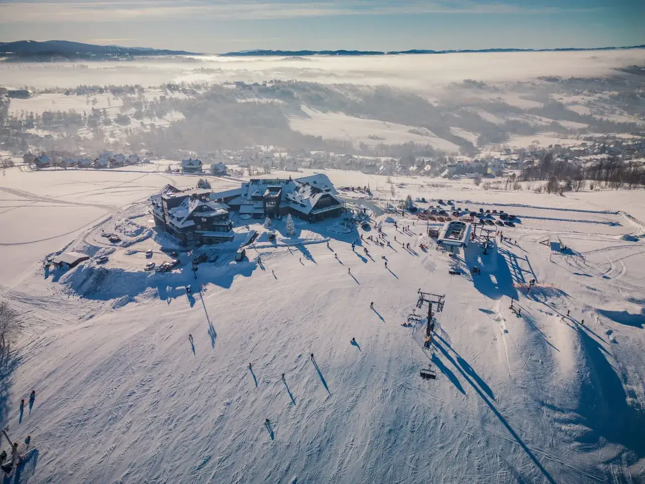 Ośnieżone stoki i hotel w Istebnej, gdzie zimą czekają liczne atrakcje. Narciarze i snowboardziści cieszą się jazdą, a kolejka linowa wwozi ich na górę.
