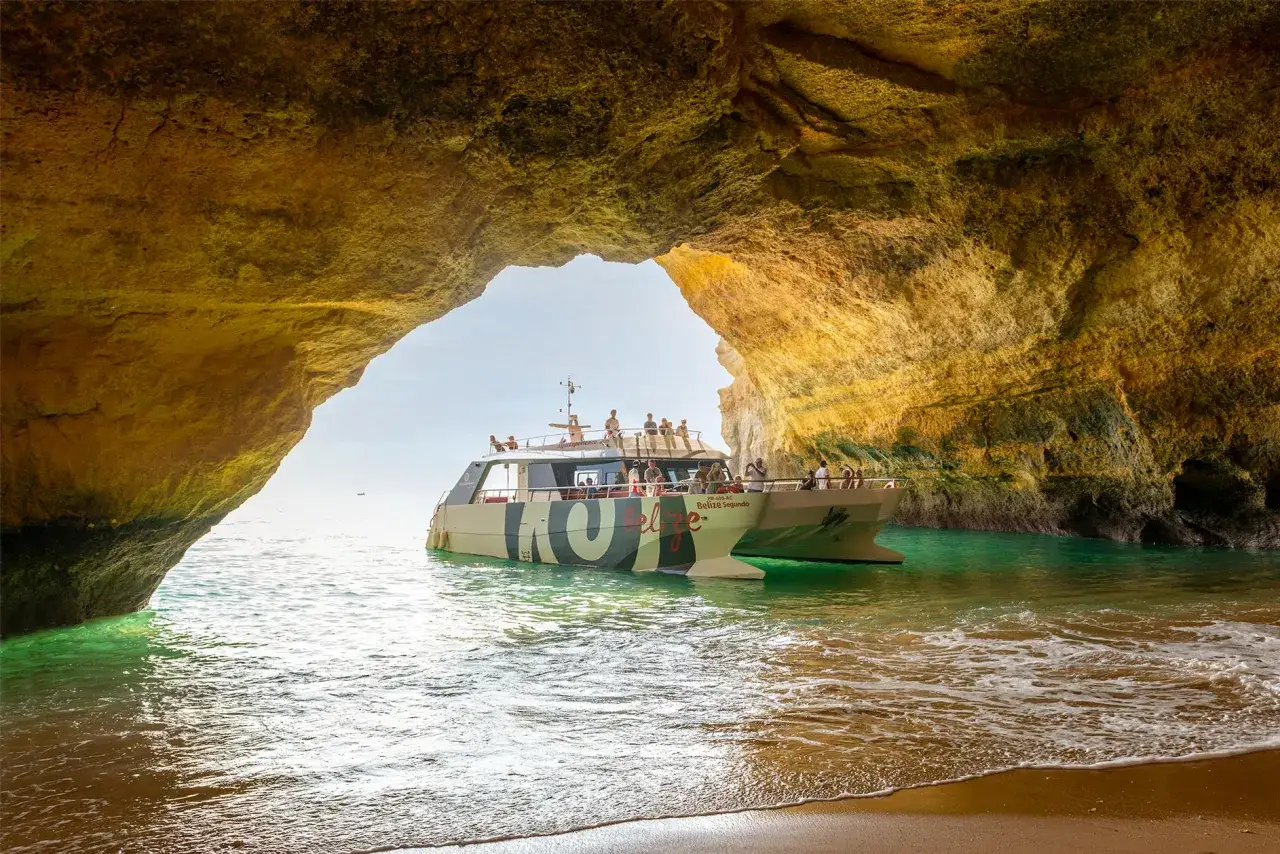 Boot mit Touristen in der Benagil-H&ouml;hle, umgeben von goldenen Felsformationen und t&uuml;rkisfarbenem Wasser.