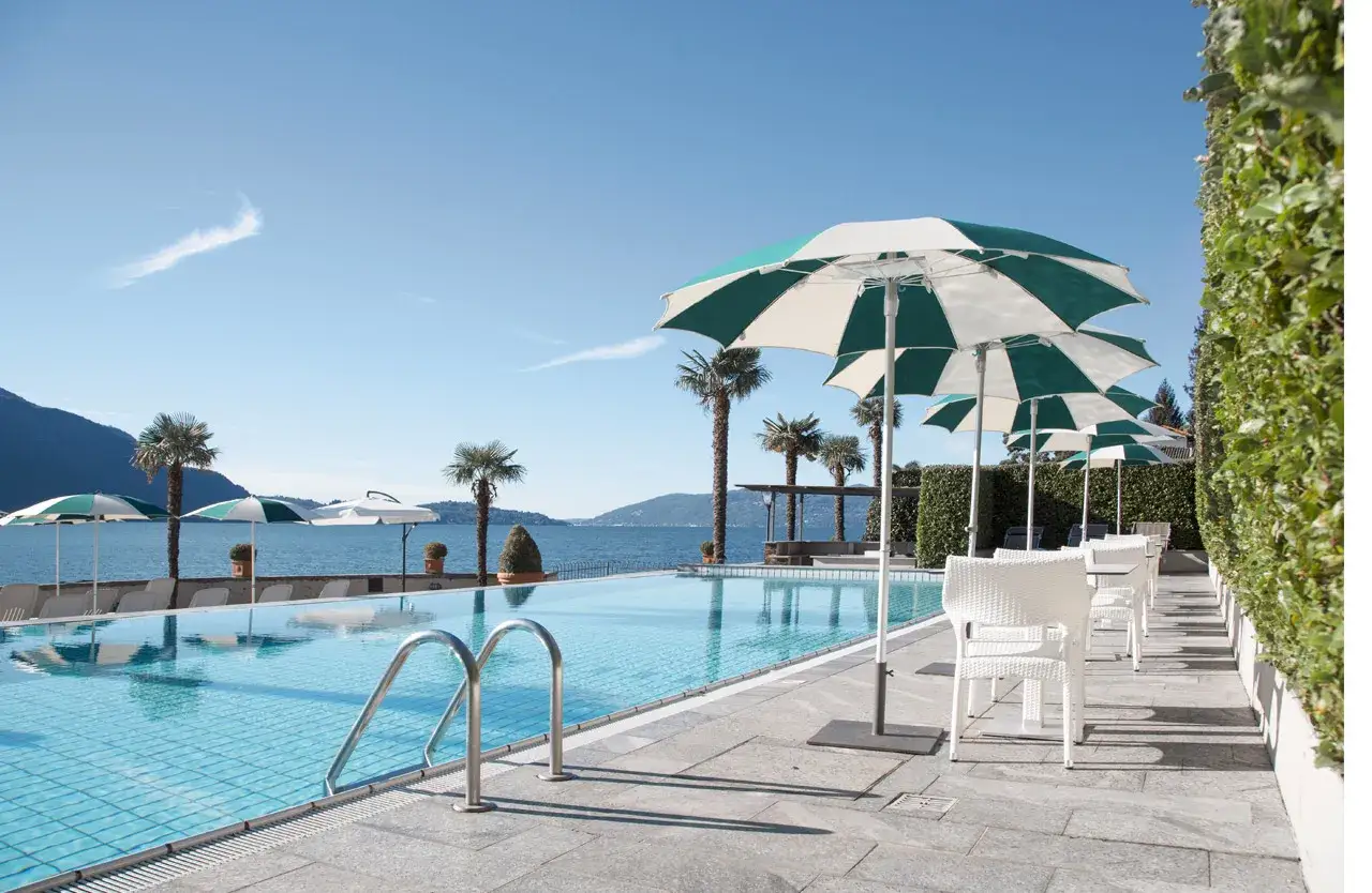 Piscine d'un hôtel à Ghiffa sur le Lac Majeur, avec parasols, palmiers et vue sur le lac.