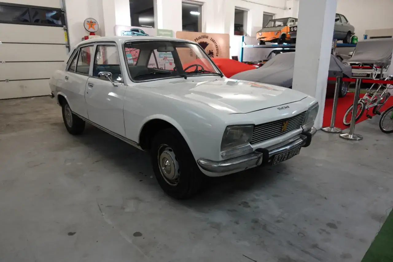 Biały Peugeot 504, klasyk z lat 70., stoi w garażu obok innych zabytkowych pojazdów.