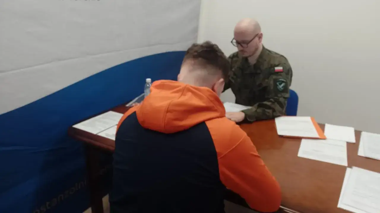 Młody mężczyzna w bluzie z kapturem siedzi przy biurku naprzeciwko żołnierza. Rozmowa dotyczy wieku poborowego w Polsce.