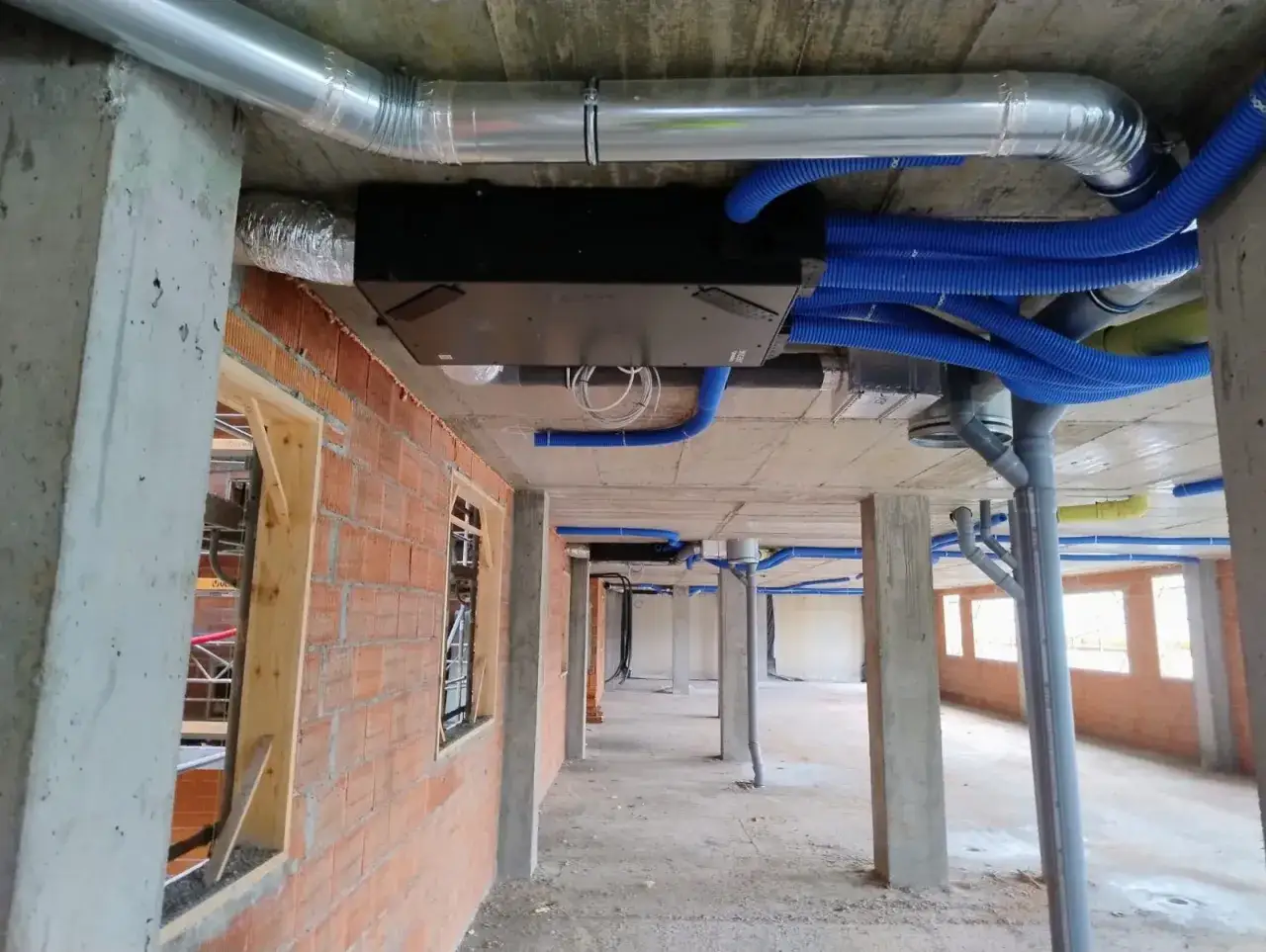 Instalaci&oacute;n de un recuperador de aire con conductos azules y grises en un edificio en construcci&oacute;n.