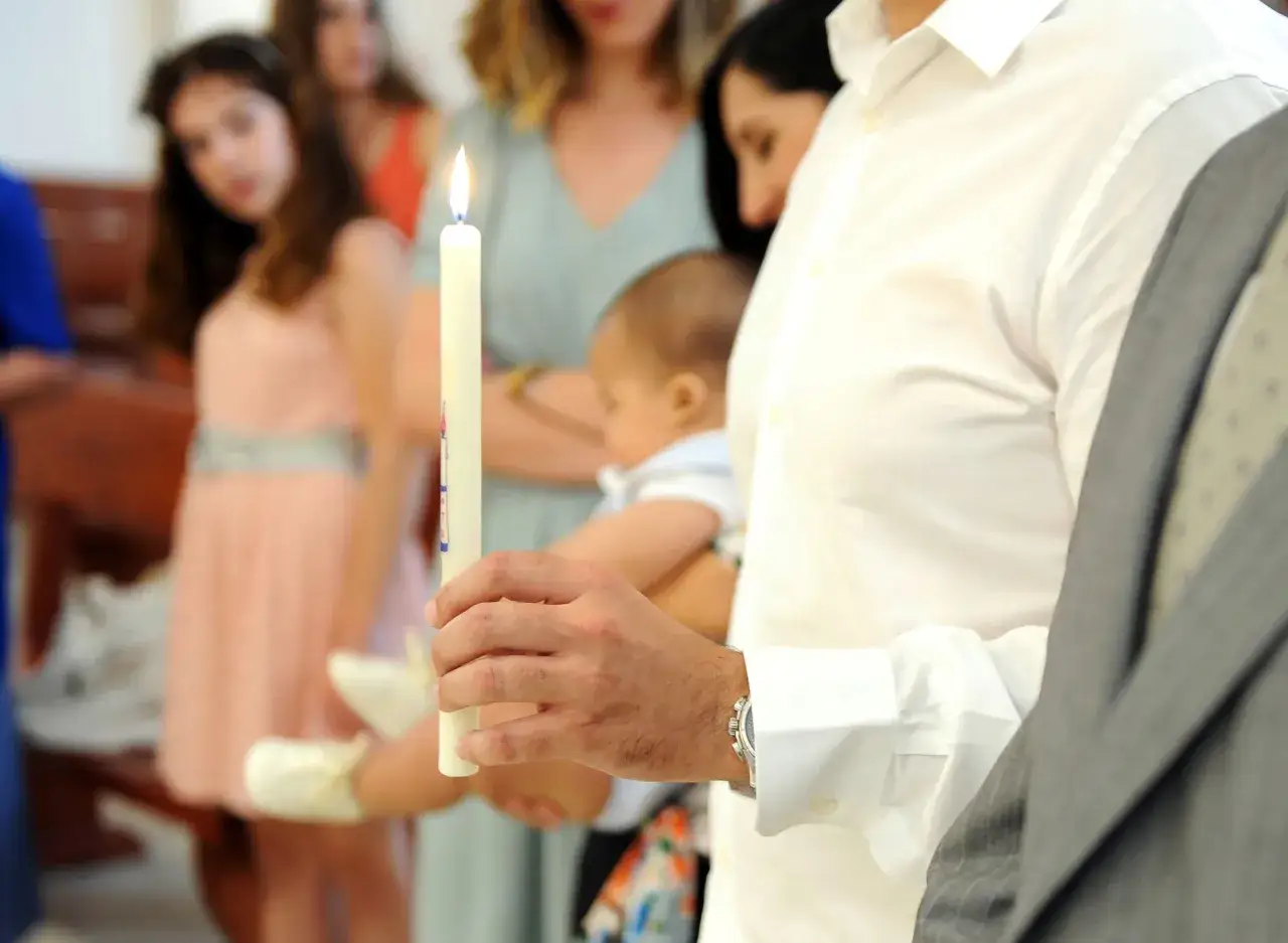 Un homme tient une bougie allumée, témoin de baptême, près d'un bébé. Des femmes et une jeune fille sont en arrière-plan.