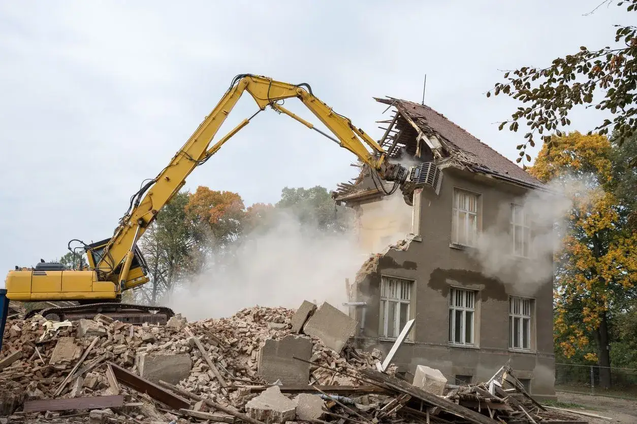 Żółta koparka burzy dom, tworząc chmurę pyłu. Rozbiórka domu kosztuje, a gruz piętrzy się u podstawy.