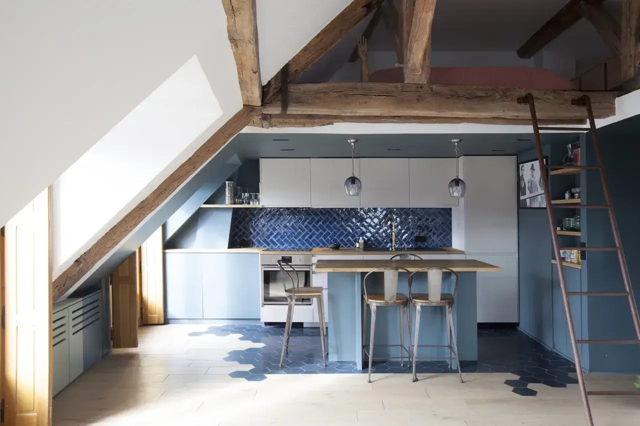 Cuisine moderne sous une **mezzanine sous toit en pente**, avec poutres apparentes, îlot central et carrelage hexagonal.