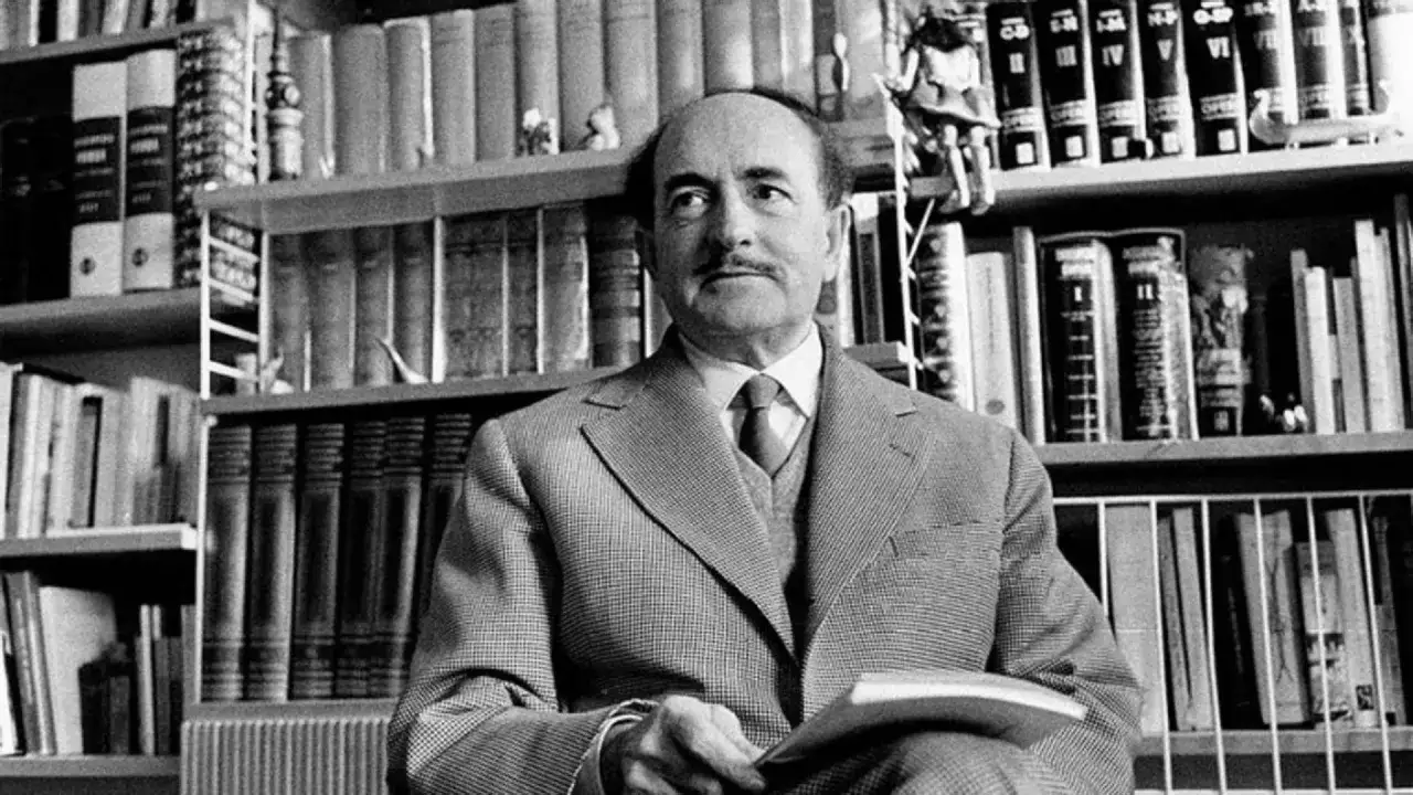 Uomo con baffi in giacca e cravatta, circondato da libri. Un'atmosfera che evoca 