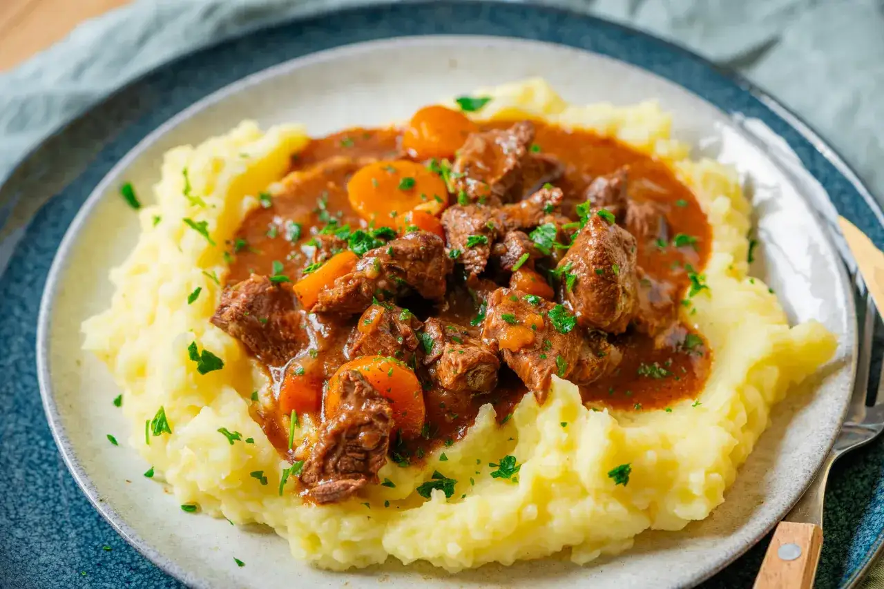Zartes, im Topf gegartes Fleisch in kr&auml;ftiger Sauce mit Karotten, serviert auf cremigem Kartoffelbrei, garniert mit Petersilie.