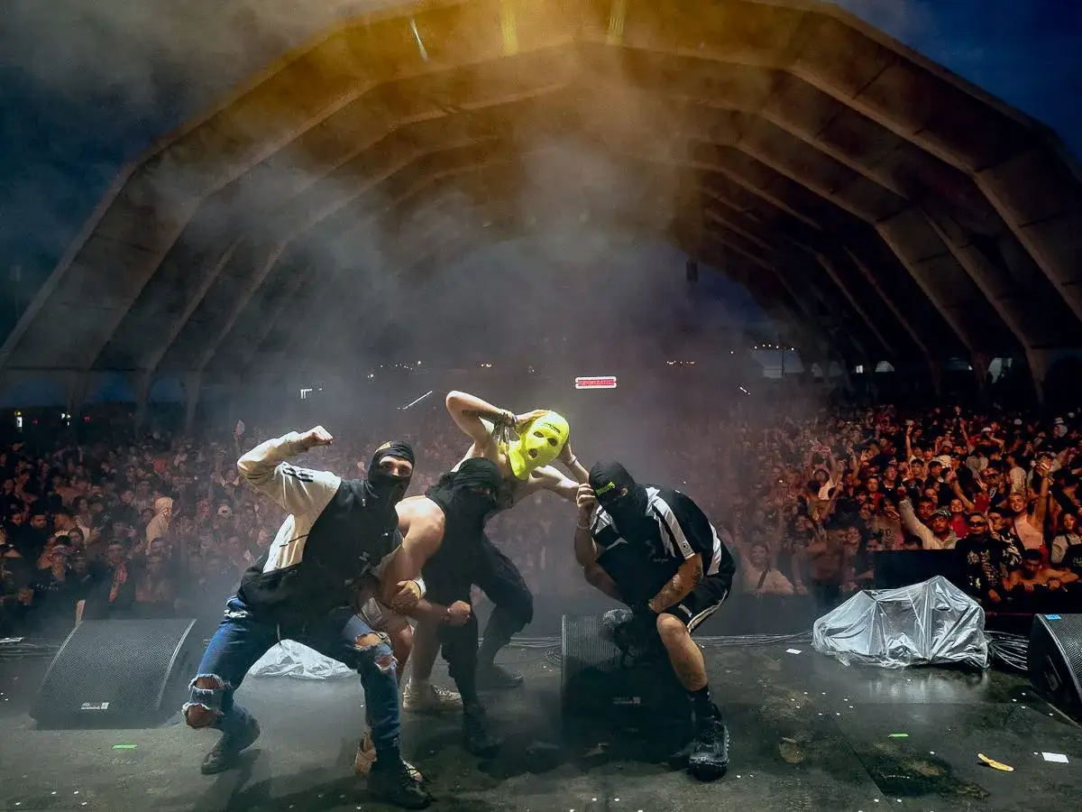Artistas enmascarados actúan ante una multitud en un concierto. El público parece vibrar con la energía, y los artistas, con sus pasamontañas y una máscara amarilla, crean un ambiente de misterio y emoción.