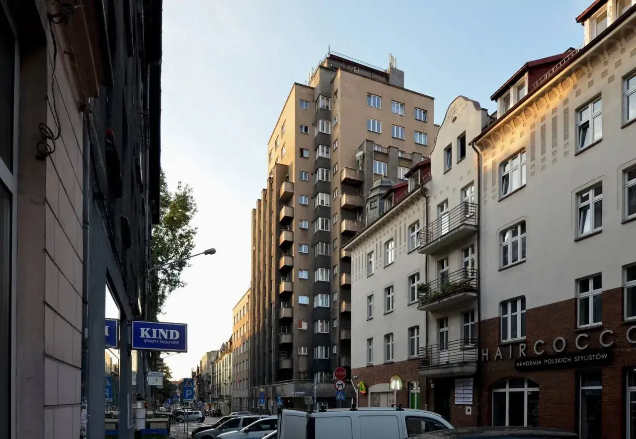 Wysoki, szary drapacz chmur w Katowicach góruje nad ulicą pełną kamienic.