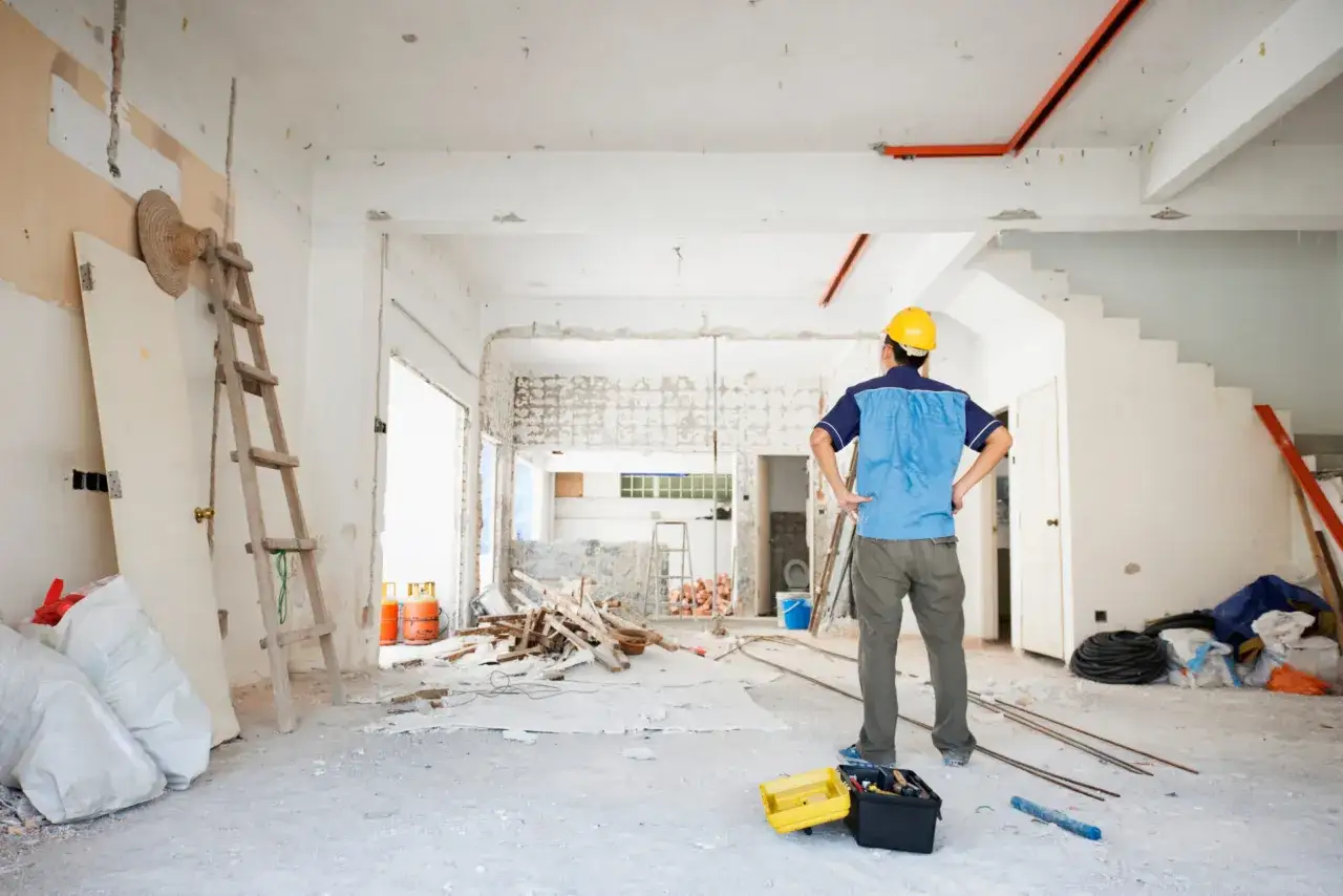 Operaio con elmetto osserva i lavori di manutenzione straordinaria in un cantiere interno, tra detriti e materiali edili.