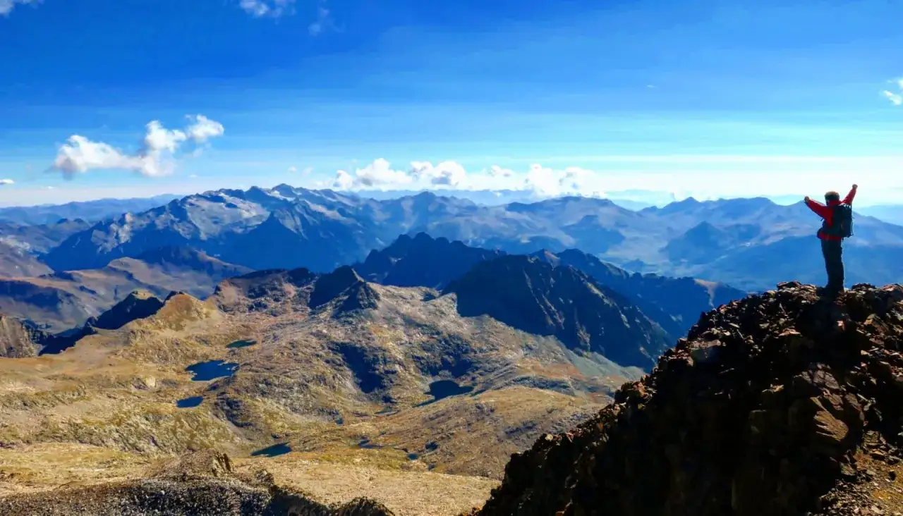 Persona celebra en la cima de un pico posets, con brazos en alto, ante un vasto paisaje monta&ntilde;oso azul bajo un cielo despejado.