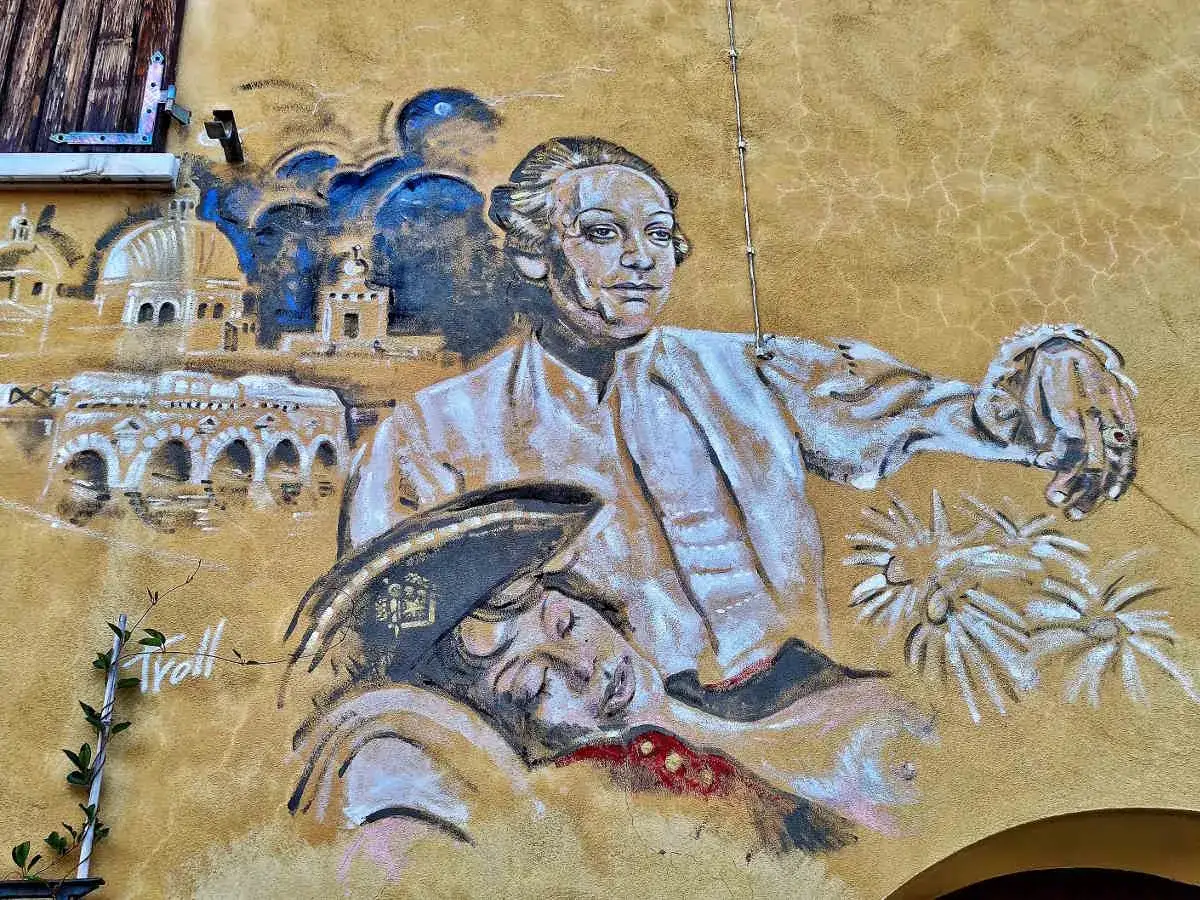 Un murale a Borgo San Giuliano raffigura un uomo con un cappello, appoggiato a una figura addormentata, con uno sfondo di architetture storiche.