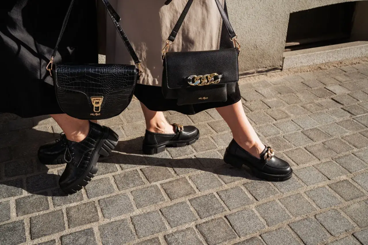 Dwie kobiety w czarnych, stylowych butach i torebkach Nine West spacerują po bruku.