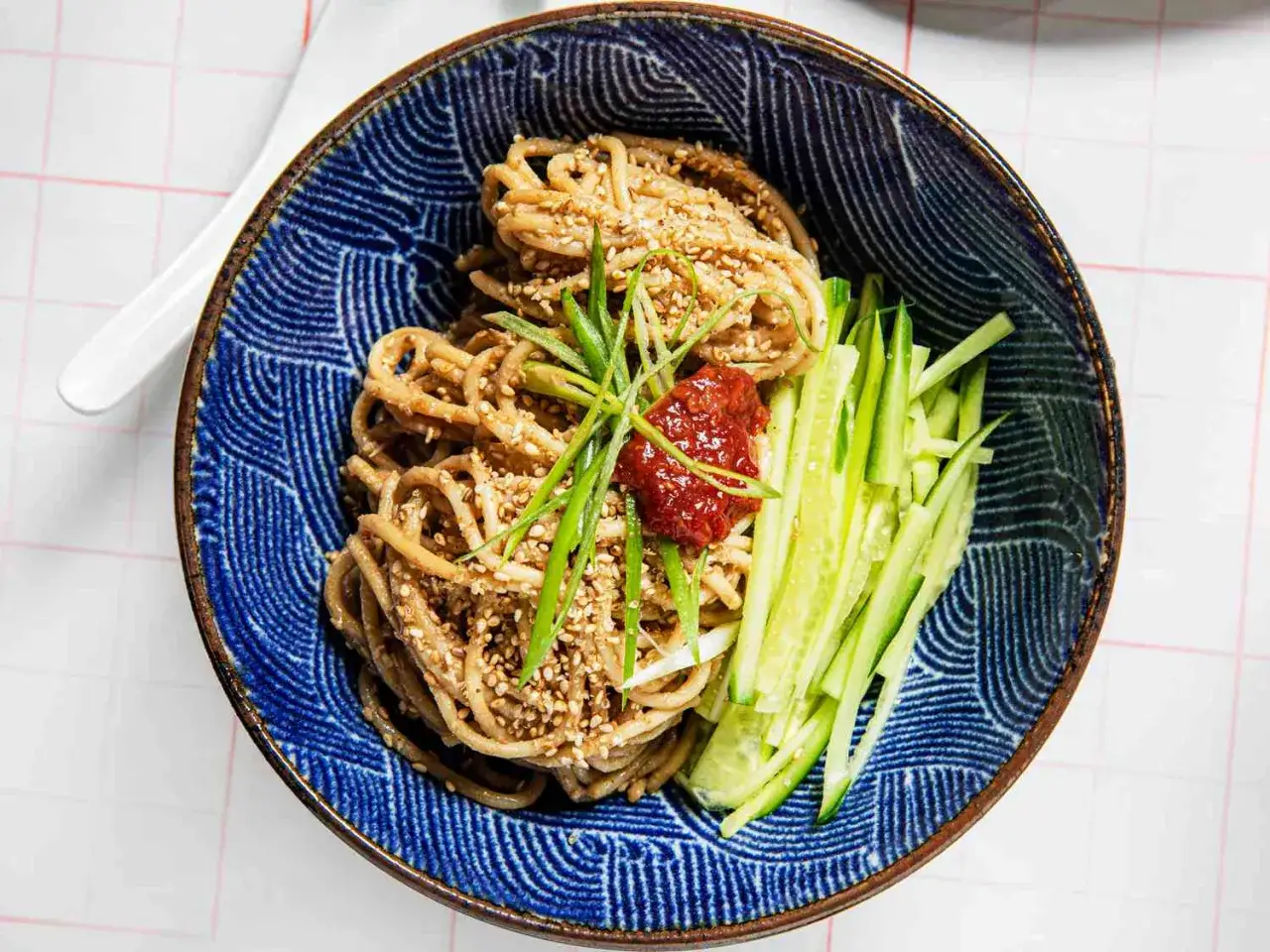 En skål med asiatisk nudelsallad med gurka, sesamfrön och en klick chilisås.