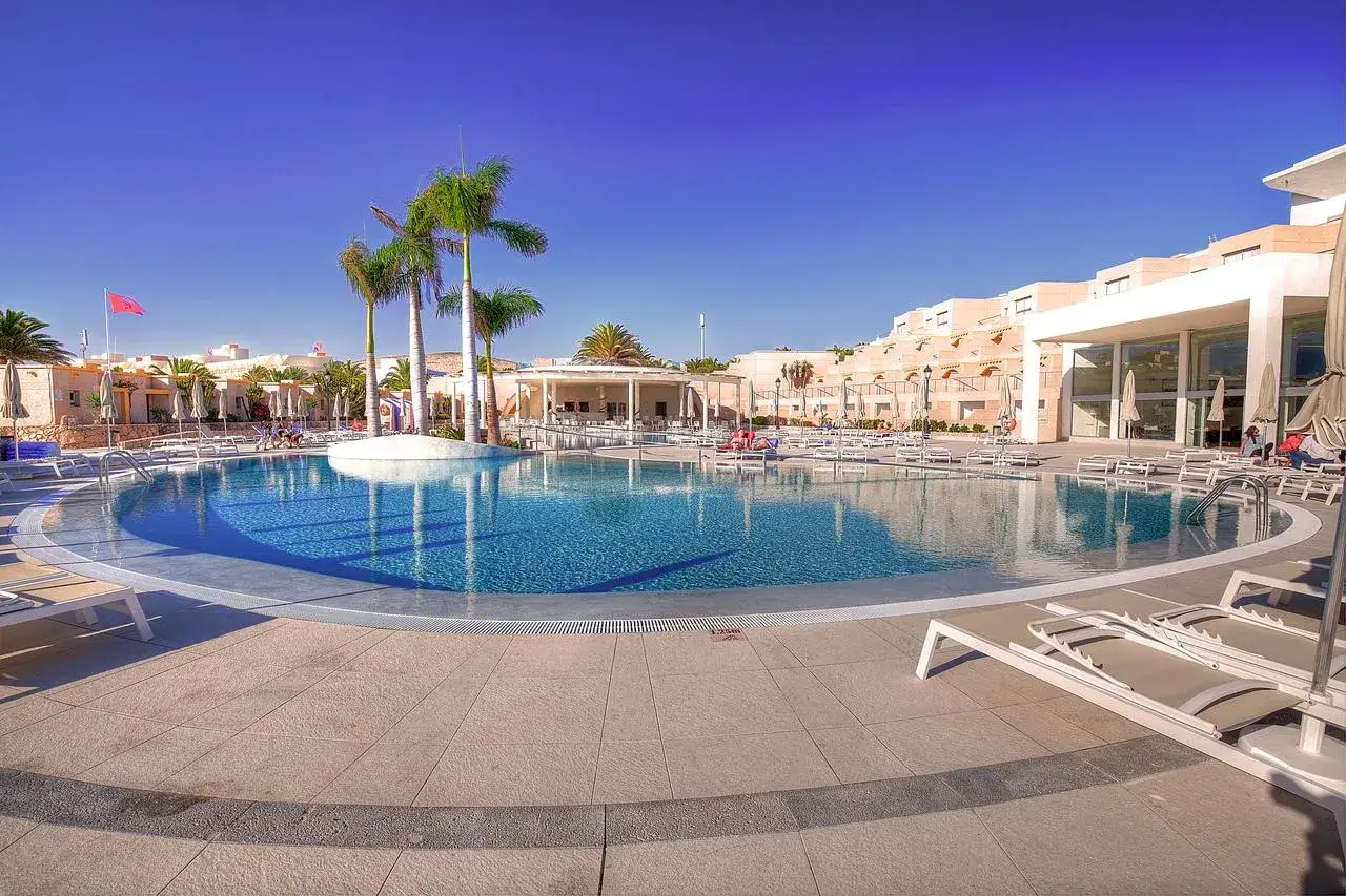 Einladender Poolbereich im SBH Monica Beach Resort mit Palmen, Liegen und einem hellblauen Himmel.