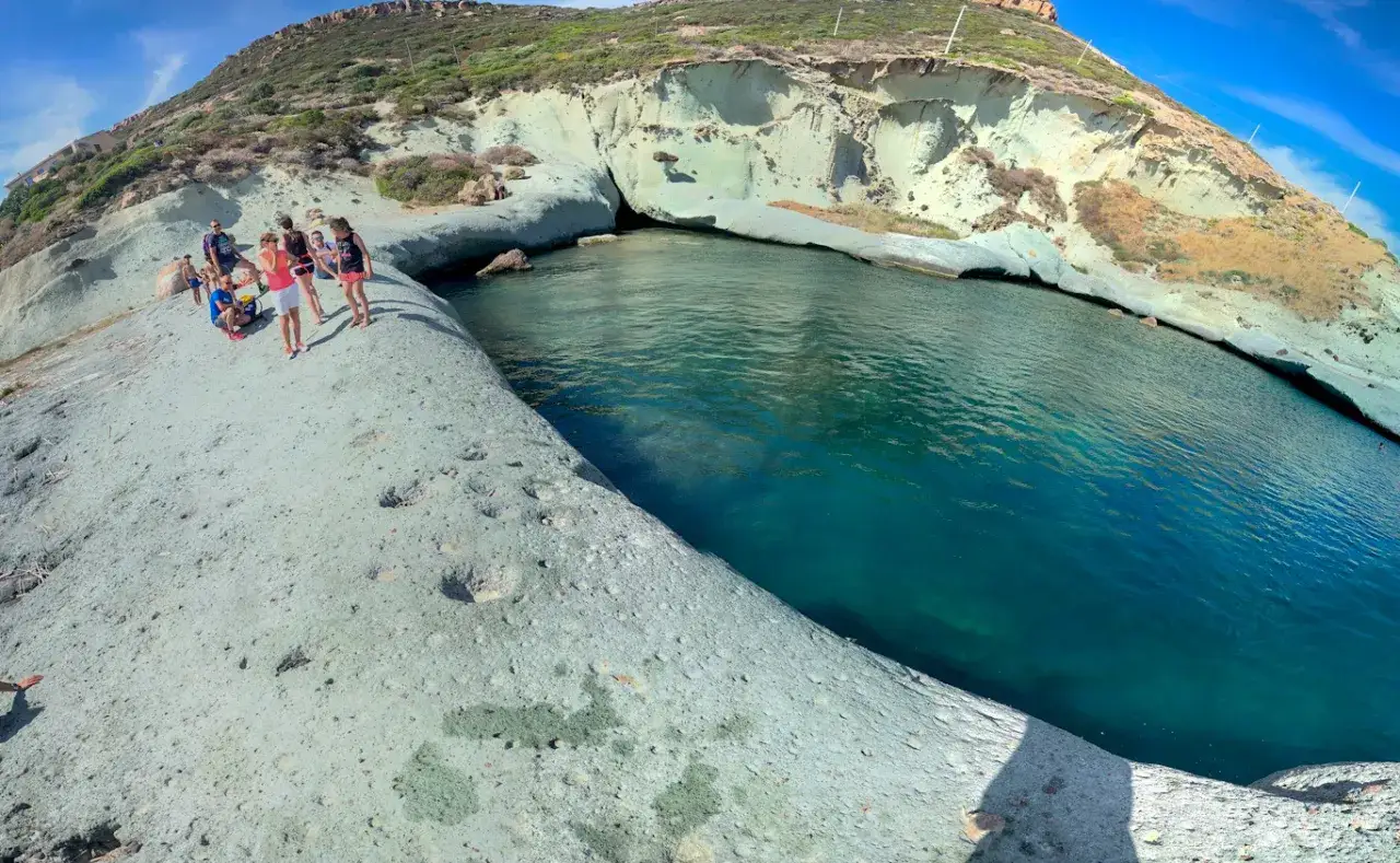 Un gruppo di persone ammira un mare cristallino da una scogliera. Un cane malu, forse un cucciolo, corre felice sulla roccia.