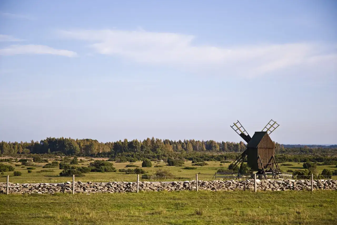 En gammal väderkvarn står på lilla alvaret, omgiven av en stenmur och grönskande landskap under en blå himmel.