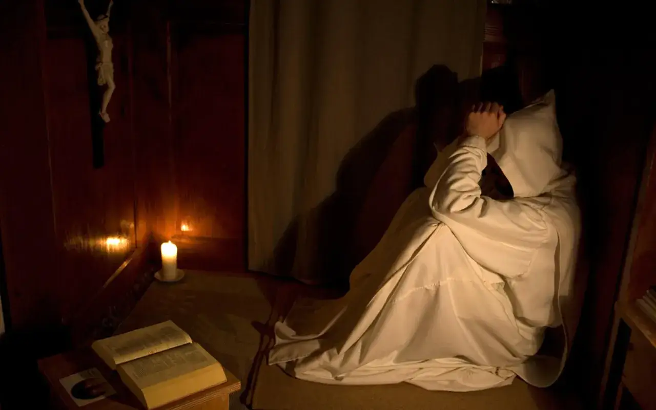 A monk in a white robe kneels in prayer during a christian silent retreat, illuminated by candlelight. A crucifix hangs on the wall.