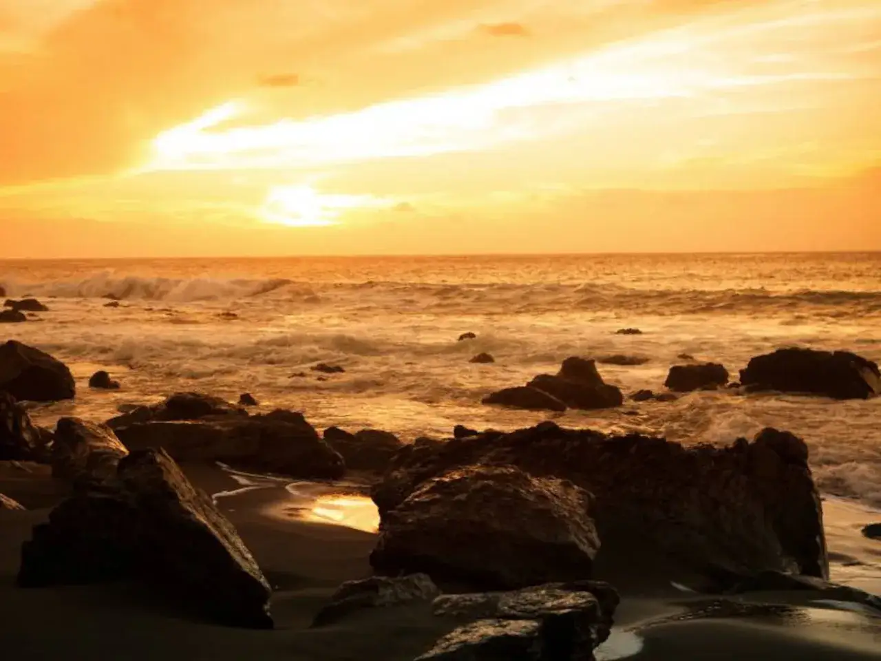 Atardecer dorado en Gran Canaria que ver: rocas oscuras en la playa y el mar con olas rompiendo bajo un cielo anaranjado.