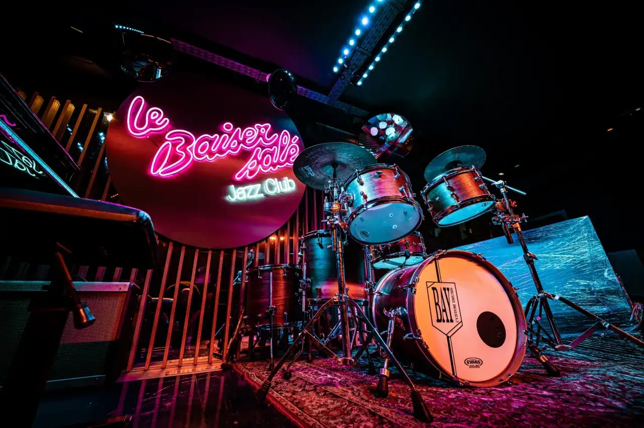 Batterie de jazz dans un club parisien, éclairée par des néons roses et bleus. Ambiance feutrée pour une soirée musicale.
