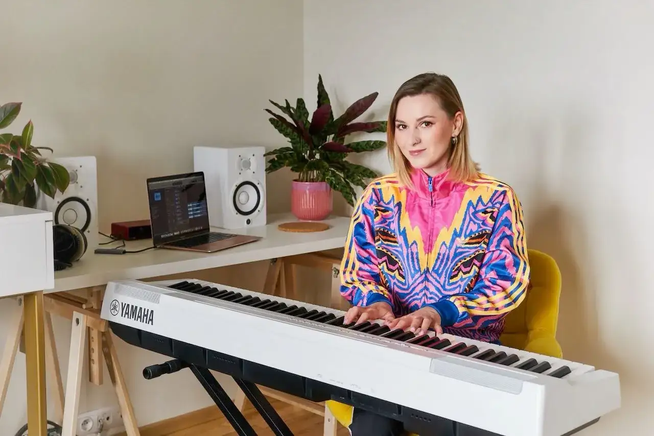 Kobieta w kolorowej kurtce gra na pianinie Yamaha. W tle laptop i głośniki, idealne do lekcji śpiewu i tworzenia muzyki.
