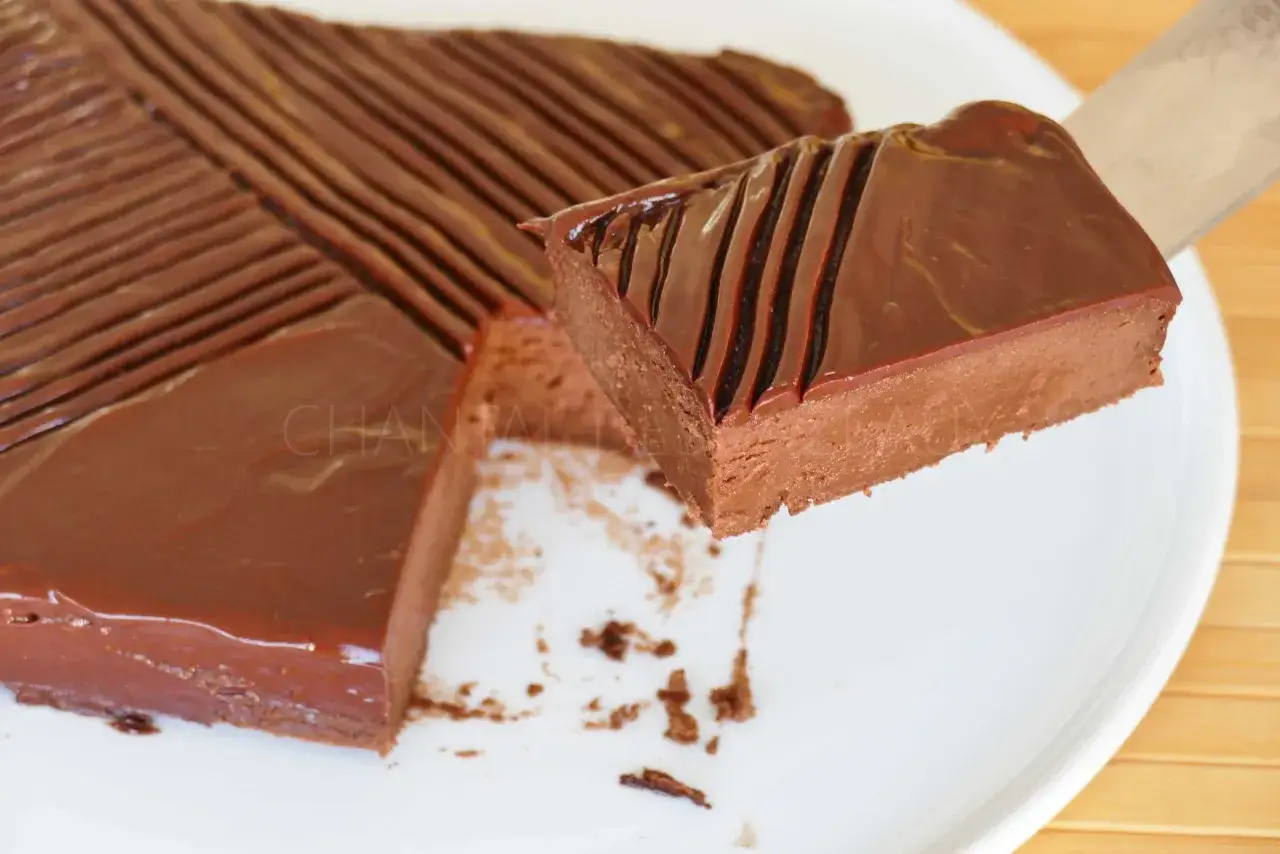 Gâteau au chocolat onctueux, décoré de lignes de chocolat noir. Une part est soulevée, révélant sa texture riche, évoquant la douceur de la chantilly mascarpone chocolat.
