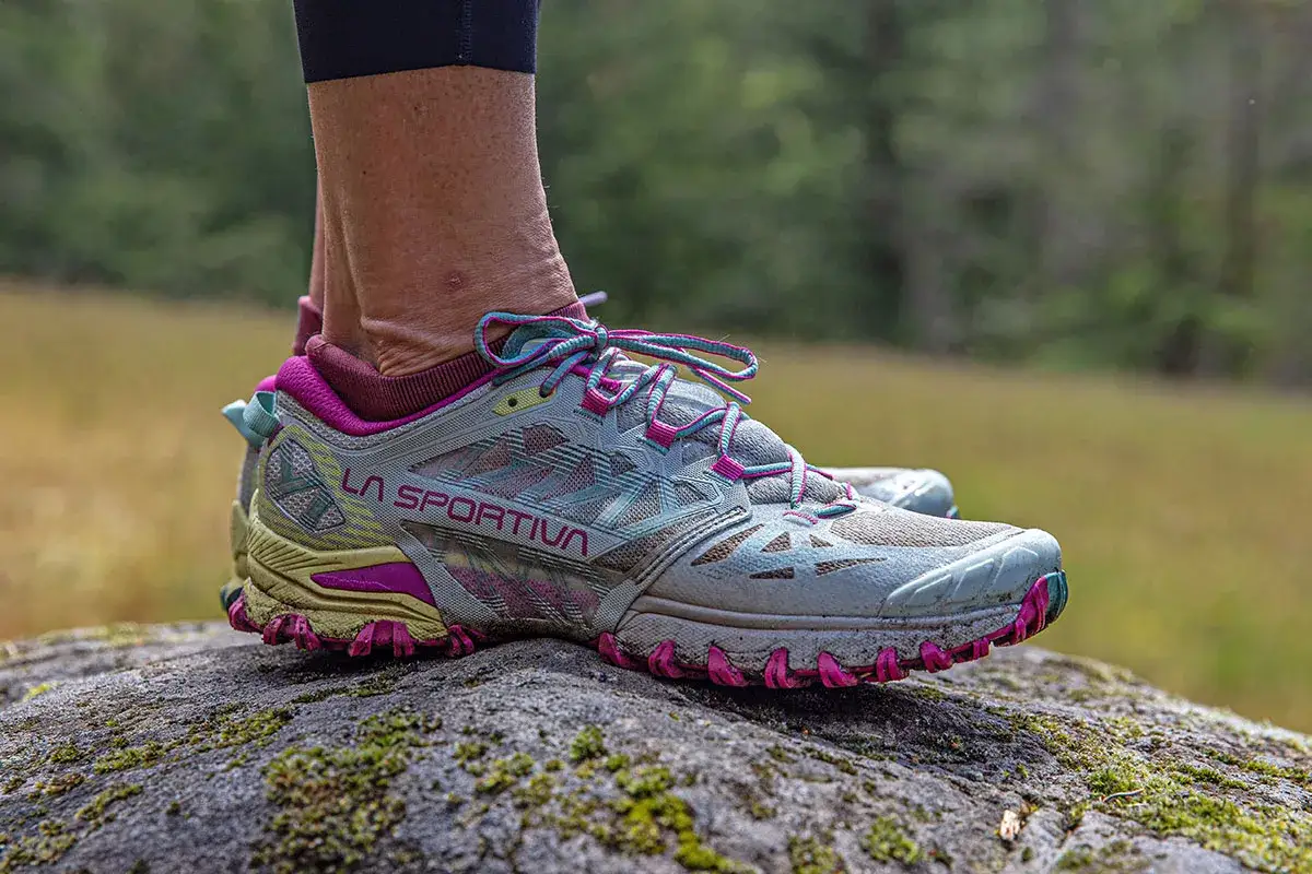 A close-up of a La Sportiva Bushido III trail running shoe on a mossy rock. The shoe features a light grey upper with pink and teal accents.