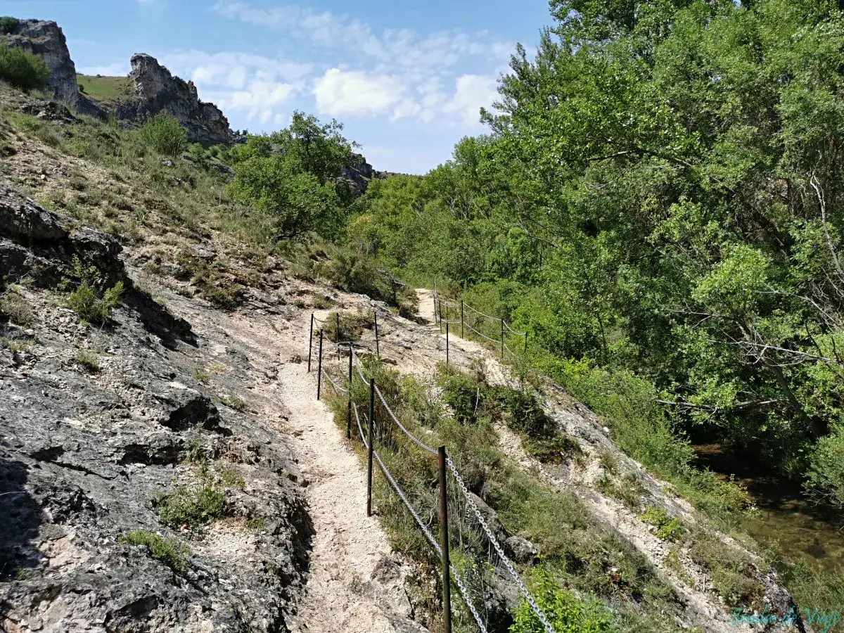 Sendero pedregoso con barandilla metálica que serpentea por la ladera rocosa del barranco del Río Dulce, bordeado de vegetación frondosa y un río al fondo.