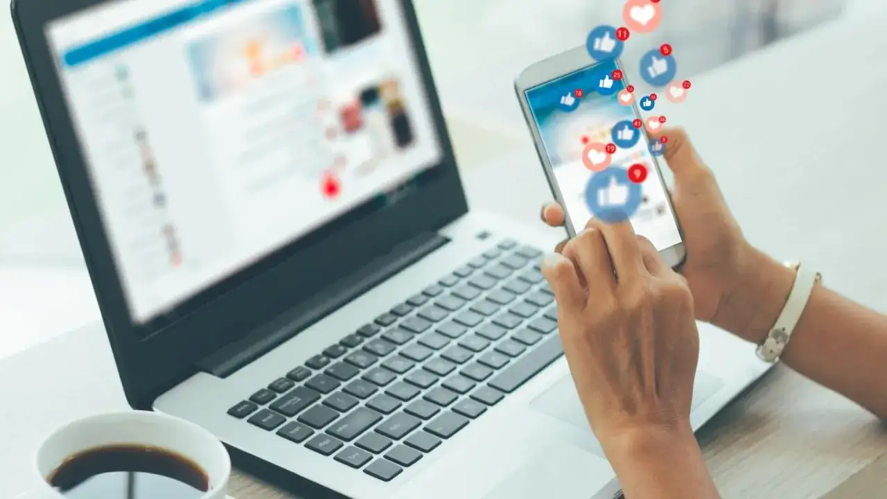 Mani che scorrono su uno smartphone con notifiche social, accanto a un laptop. Il lavoro di un social media manager stipendio è legato a queste interazioni.
