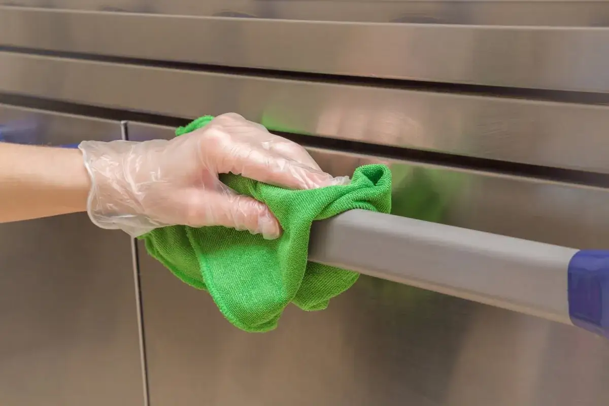Main tenant, une main gantée utilise un chiffon vert pour nettoyer une barre en inox. Le bicarbonate de soude est idéal pour nettoyer l'inox.
