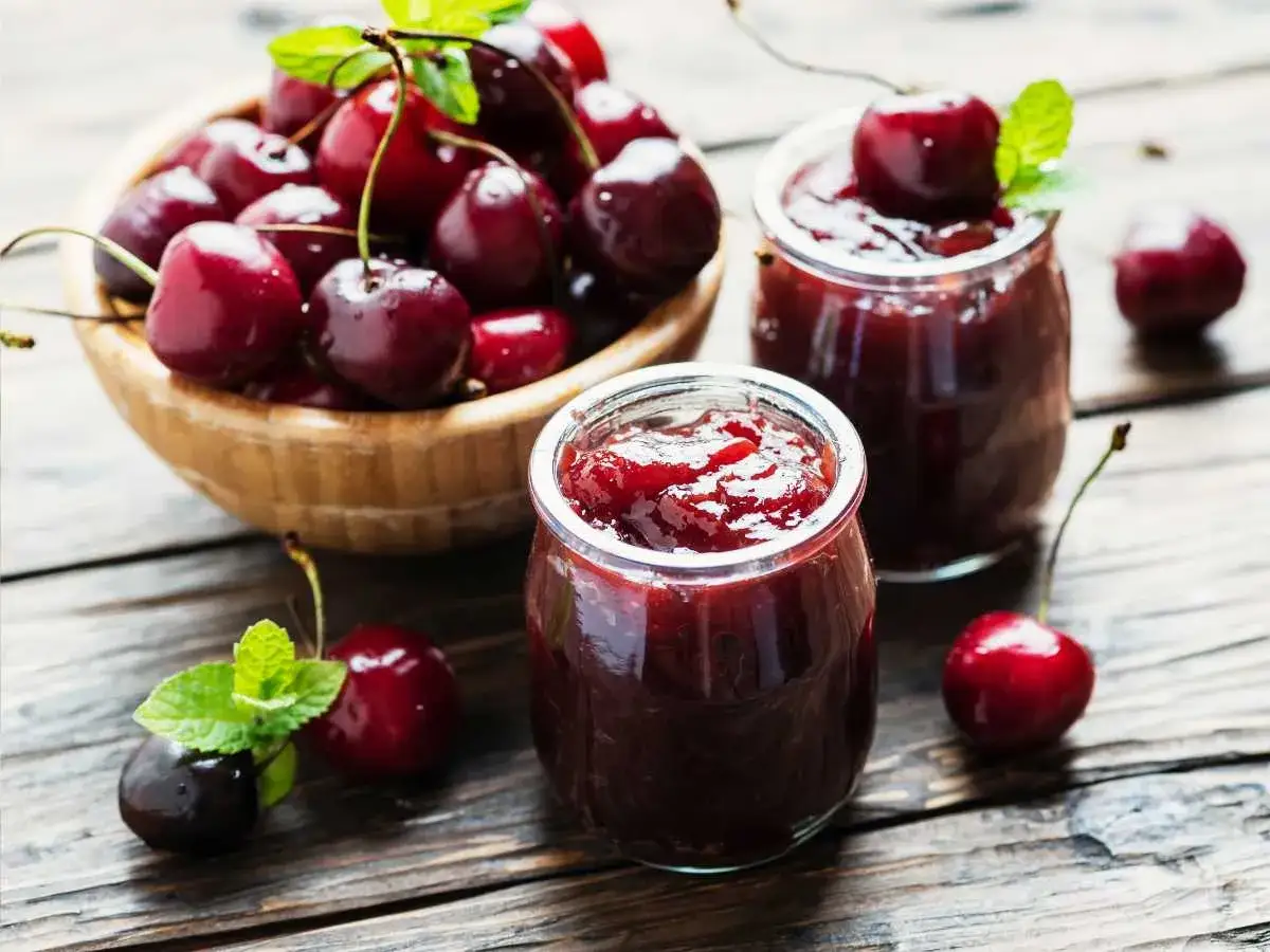 Dos frascos de mermelada de cerezas casera, con cerezas frescas y hojas de menta sobre una mesa de madera r&uacute;stica.