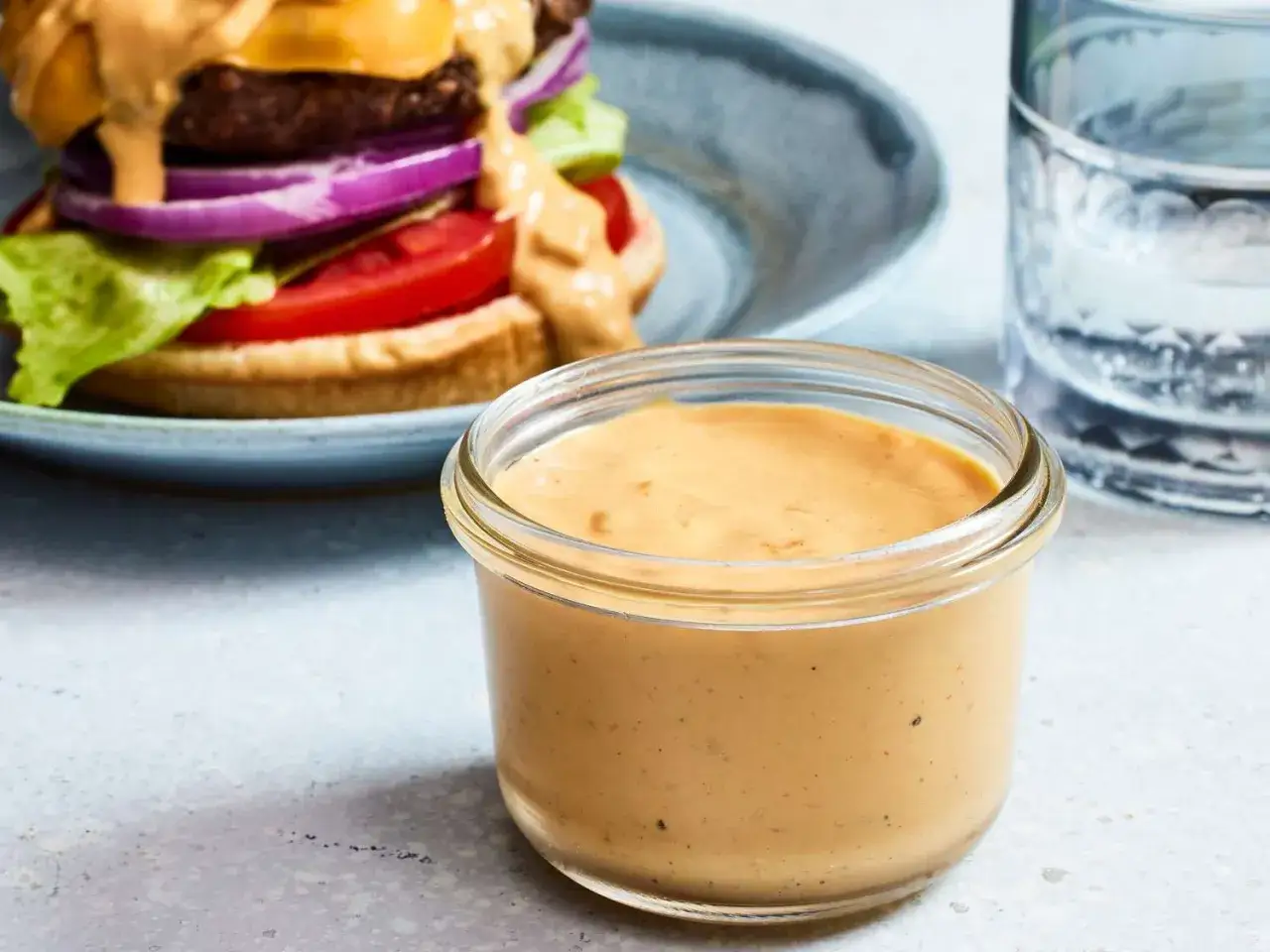 Ein Glas mit cremiger Burger-Sauce, perfekt für dein nächstes Burger-Rezept. Im Hintergrund ein saftiger Burger mit Käse, Tomate und Zwiebeln.